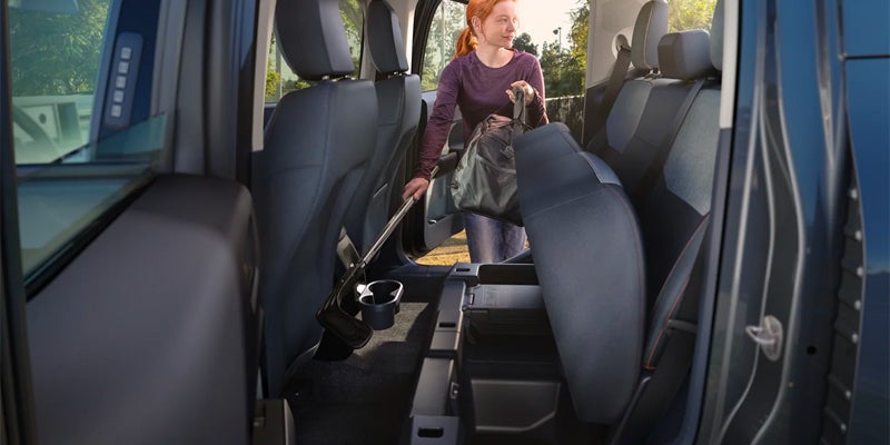 A woman installs a portable vacuum cleaner with a long handle and a large bag into the back of a car.