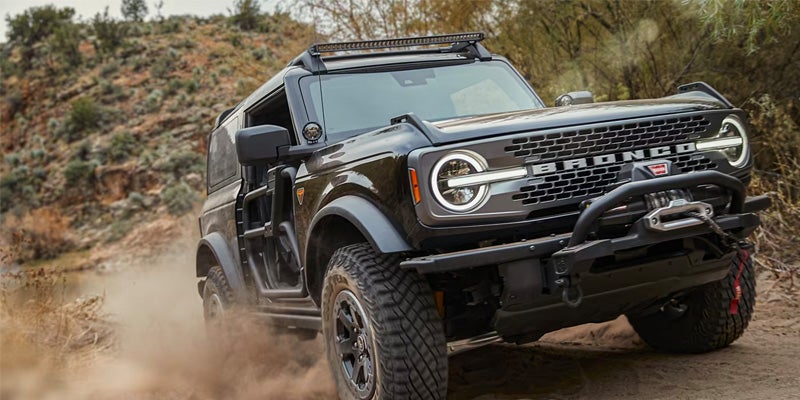 A black two-door Ford Bronco with a winch and roof-mounted light bar kicks up dust on a dirt trail.