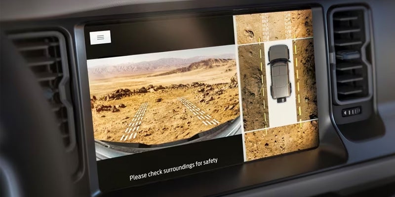Vehicle infotainment screen displaying a desert landscape from three camera angles, with car icon and guide lines.