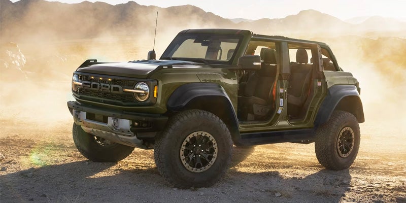 An army green Ford Bronco Raptor with no doors or roof is parked on an unpaved road with mountains in the dusty distance.