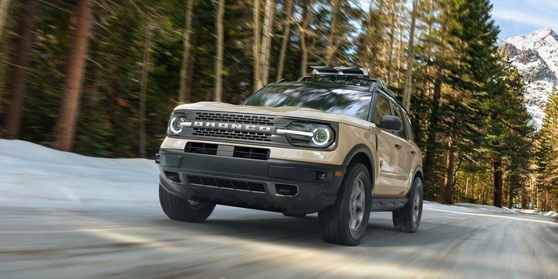 A tan Ford Bronco Sport drives on a snow-dusted road through a forest with a snow-capped mountain in the background.
