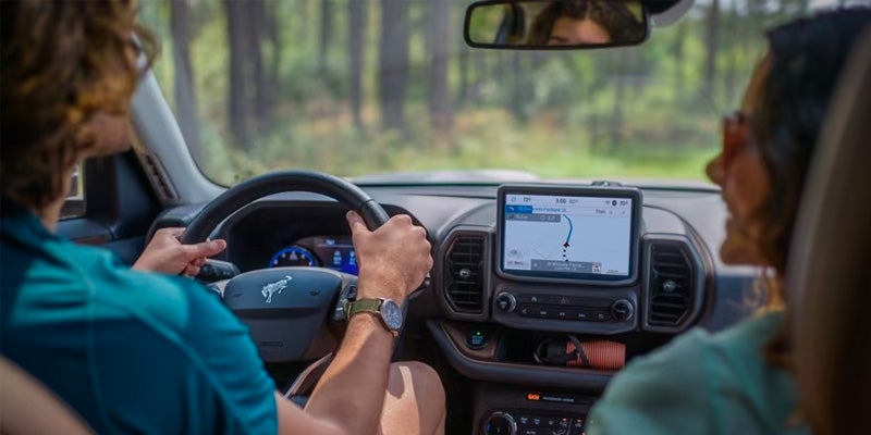 Inside a car, a person drives with a navigation screen showing a route, while a passenger glances to the side.