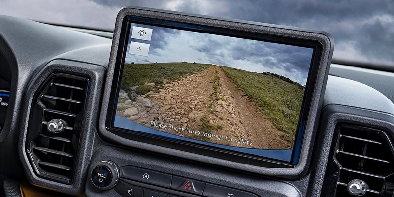 In-car display showing a rocky, unpaved off-road trail curving uphill over grass under a stormy sky.