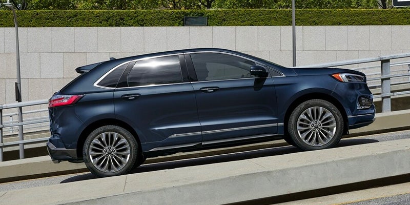Side view of a dark blue SUV parked on an incline, with a concrete wall and green hedges behind it.
