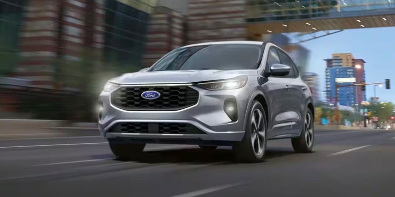 A silver Ford Escape drives on a city street, headlights on, with blurred buildings in the background.