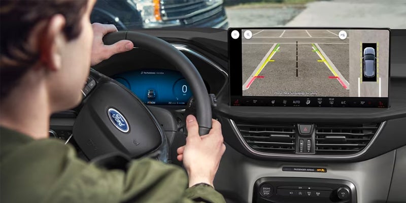 Driver's hands on a Ford steering wheel, an instrument cluster display reads 0, and a screen displays a rearview camera with parking lines.