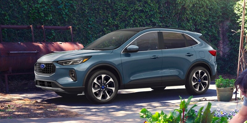A blue-green Ford Escape parked on a paved surface next to an old red smoker and a lush green wall.