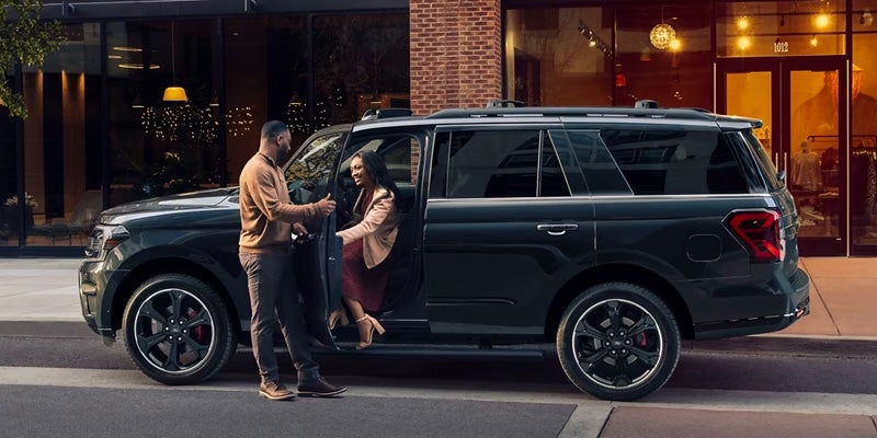 A man in a brown sweater and dark pants helps a woman in a red dress and tan jacket into a dark SUV at night.