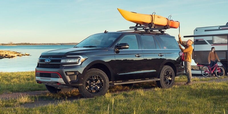 Black Ford Expedition with a kayak on its roof, parked by a lake, with people and a trailer nearby.
