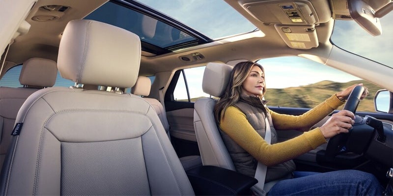 A woman in a vest, gray shirt, and jeans driving a car with a panoramic sunroof on a sunny day.