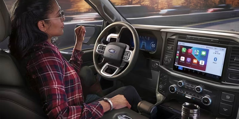 Driver's hands on a Ford steering wheel, an instrument cluster display reads 0, and a screen displays a rearview camera with parking lines.