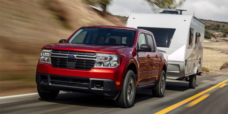 A red Ford Maverick pickup truck pulls a white travel trailer down a rural highway.