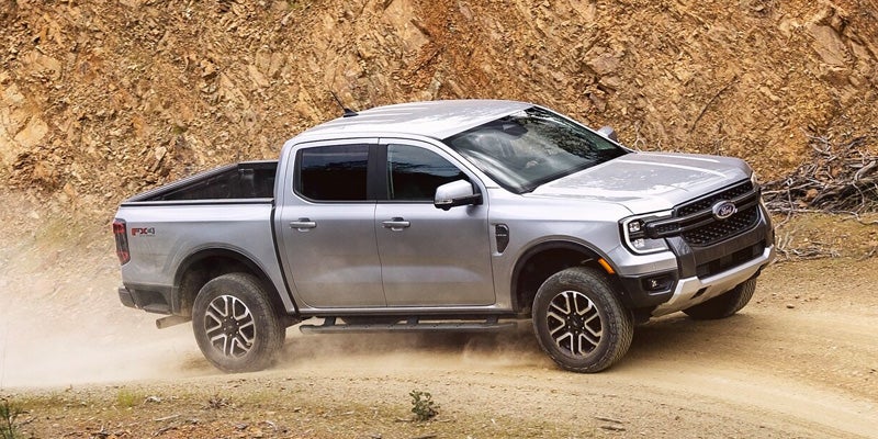 A silver 2024 Ford Ranger truck with four doors and a bed with a cover driving on a dirt road.