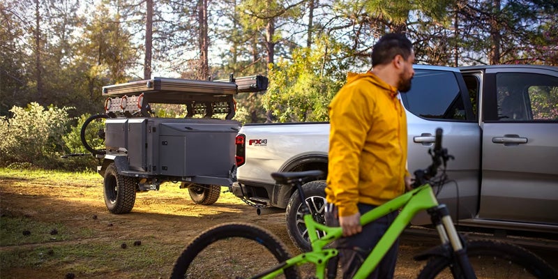 Man in yellow jacket with a green mountain bike next to silver pickup towing a gray off-road camping trailer.