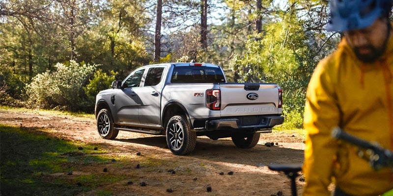 Light gray Ford Ranger pickup truck parked on a dirt path in a wooded area, with a person on a bike in the foreground.