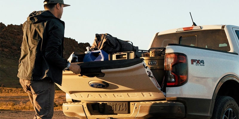 A man loading gear, including a Yeti cooler and blue canister, into the open tailgate of a beige Ford Ranger pickup truck.