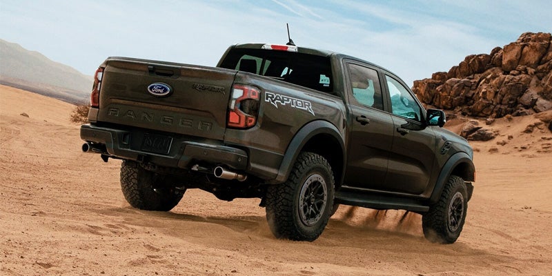 Back view of a dark grey Ford Ranger Raptor pickup truck driving through a sandy desert landscape.
