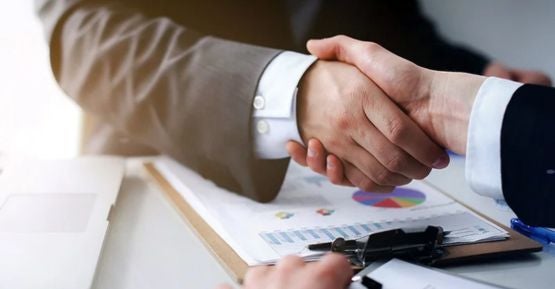 Two people in suits shaking hands over a blurred desk with papers and charts.