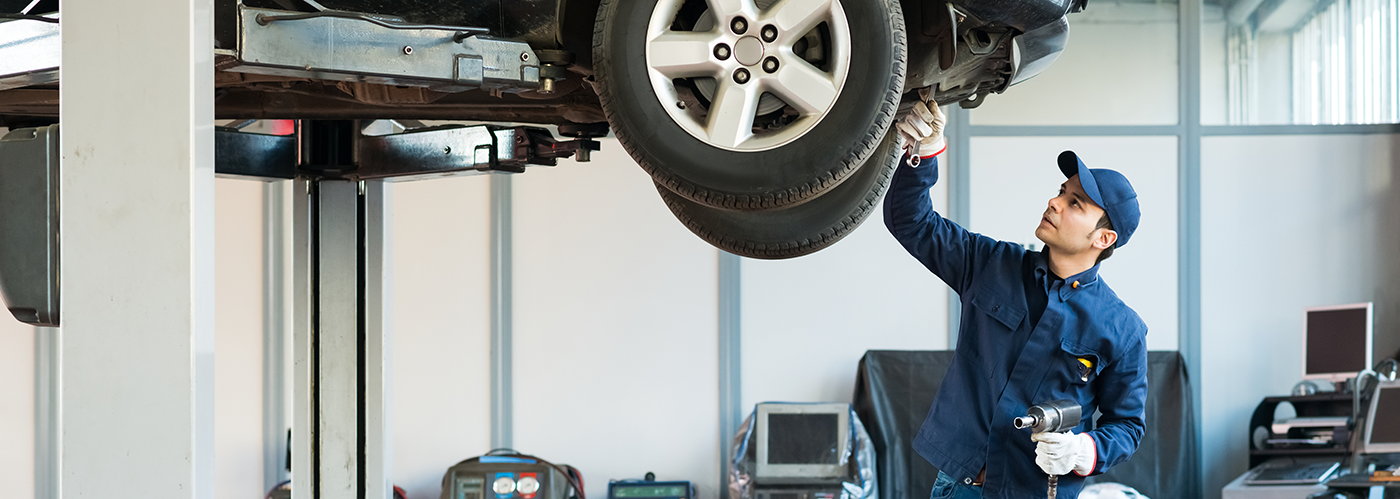 A mechanic in a blue uniform and cap inspects the underside of a car lifted on a hydraulic jack.