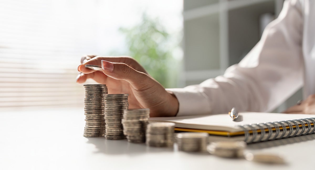 A person's hand placing a coin onto a stack, forming an ascending line of coin stacks on a desk.