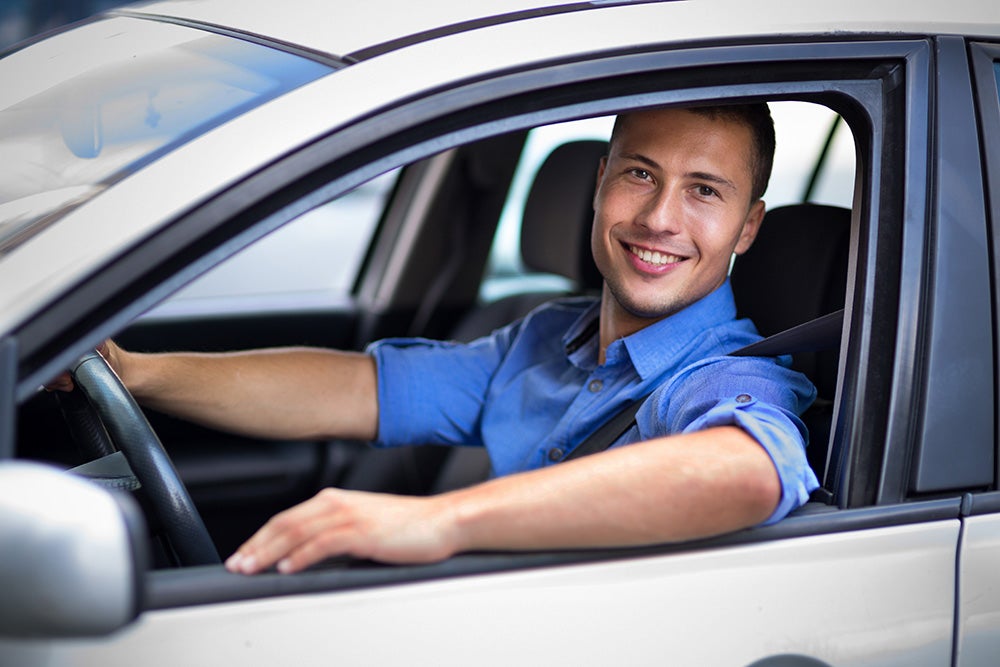 Smiling man in a blue shirt driving a white car, looking out the window with hands on the steering wheel.