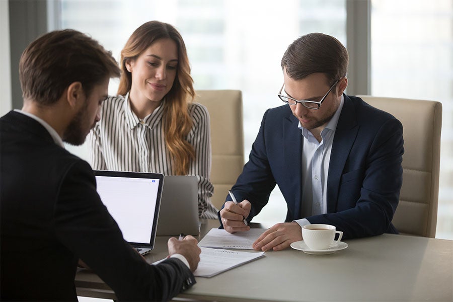 Three professionals, two men and one woman, review and sign documents at an office table.