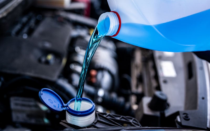 Blue windshield washer fluid being poured from a transparent bottle into a car's reservoir.