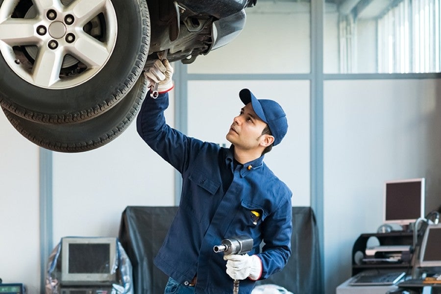 A mechanic in a blue uniform and cap inspects the underside of a car with a wrench and holds an impact gun.